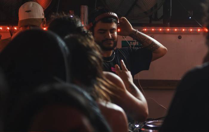 A DJ eyes closed, hand on his headphones, a string of red bulbs behind him